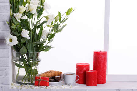 Japanese roses in a vase with water next to red candles, tea with a mint, a croissant and a cake on the windowsillの写真素材