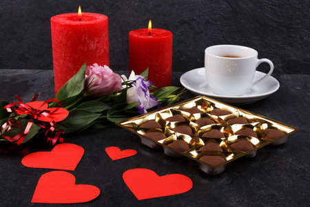 A cup of tea on a stand with a box of chocolates and burning candles, a bouquet of Japanese roses and carved hearts on a stone backgroundの写真素材