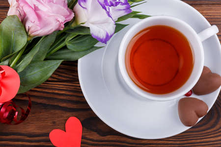 A cup of tea on a stand with candies in the form of hearts with a bouquet of Japanese roses on a wooden background. View from aboveの写真素材