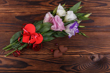 A bouquet of Japanese roses tied with a ribbon with a heart next to candy in the form of hearts and a beak lie on a wooden background. View from above...の写真素材