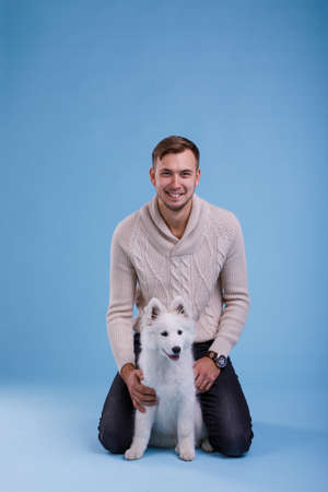 A happy guy sits and plays with a cute Samoyed puppy. On a blue background.の写真素材