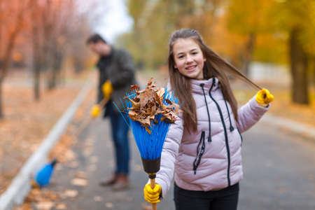 The girl is holding a broom with leaves on the back of a blurry plan dad and autumn parkの写真素材