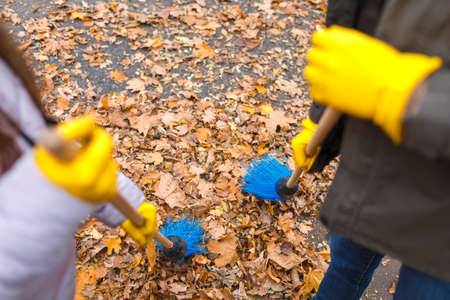 Dad and daughter in yellow prints sweep blue broom leaves on the road in the autumn parkの写真素材