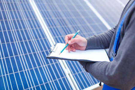 Worker in blue uniform with a tablet making notes close-up against a background of solar panelsの写真素材