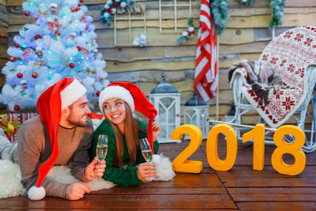 Close-up of a young couple in santa hats, holding glasses with champagne in hands, lying on the...