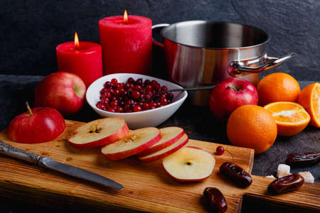 On a wooden board, juicy apples are cut, next to a metal saucepan, a deep white plate with cranberries and two burning red candles. Indoors. Side view.の写真素材