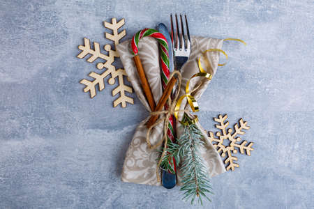 On a worn blue surface, the knife and fork are wrapped in a serving napkin and decorated with a golden ribbon, fragrant cinnamon sticks, Christmas candy and small twigs of a Christmas tree, beside wooden decorative snowflakes. View from above.の写真素材
