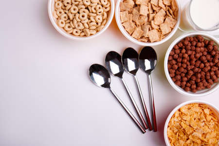 The top view of different types of cornflakes, in plates, four spoons, in the right corner is milk.