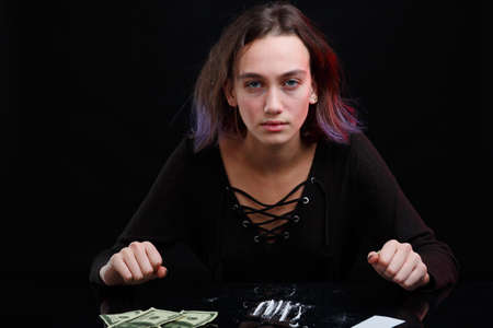 A girl drug addict sits at the table with a dosed cocaine next to the Dolar bills. On a black background.の写真素材