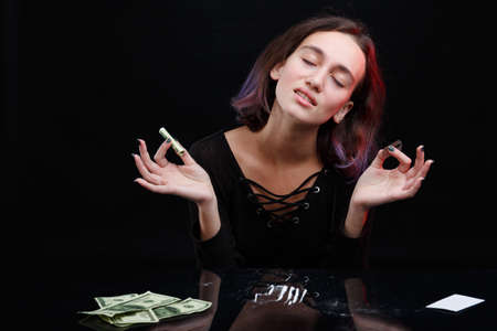 A drug-dependent girl shut her eyes and holds a twisted dollar bill and a sharp blade. On a black background.の写真素材