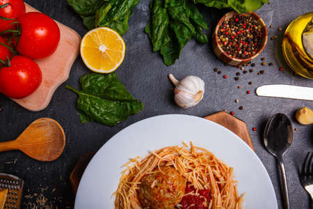 Close-up of spaghetti with grated cheese and meatballs on a plate, on a wooden background, on the table is garlic, green spinach, tomatoes, cutlery, pepper sprinkled with peasの写真素材