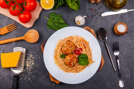 Close-up of spaghetti with grated cheese and meatballs on a plate, on a wooden background, on the table is garlic, green spinach, tomatoes, cutlery, pepper sprinkled with peasの写真素材