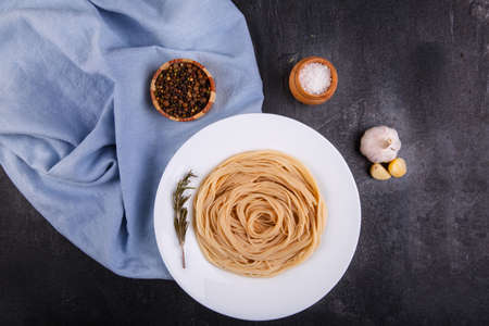 Spaghetti of hard varieties in a white plate, with a sprig of marble, on dark concrete, next to it lies garlic, salt, and picking peppers in peas, a plate stands on a blue fabric. View from above.の写真素材