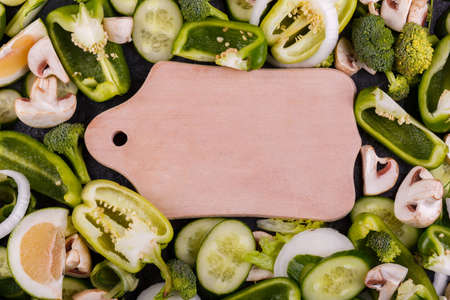 Close-up vegetables, broccoli, bell pepper, pieces of cucumber, champignons, onion rings, and lime are planted, in the middle of vegetables lies a wooden cutting board, the concept of a healthy dietの写真素材