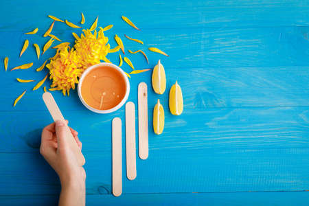 The hand holds a wooden stick next to the wax in a cup, chrysanthemums, wooden sticks and lemon slices on the left on a blue wooden backgroundの写真素材