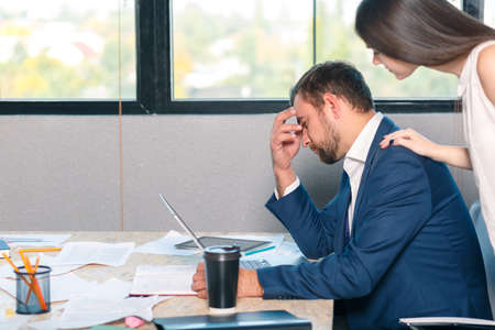 A sad man sits at the desk and clings to his head, an employee stands beside him and comforts him.の写真素材