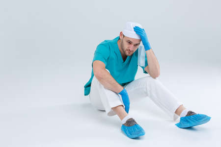 A frustrated doctor in uniform, sits on the floor and nervously holds one hand by the head. Isolation.の写真素材
