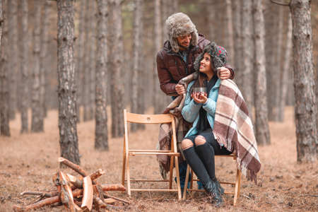 A couple in love in forest, a girl is drinking tea, a guy hides a girl with a warm blanket from behind.の写真素材