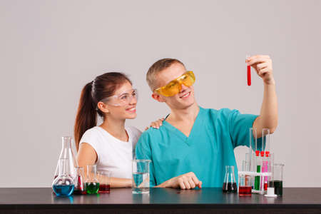 A pair assistants, holding a glass flask with a red liquid and looking at it. Indoors laboratory....