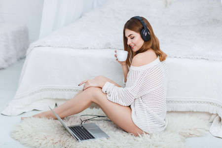 Young girl sitting on the floor , looking at laptop in headphones, smiling and holding a cup. Indoors.の写真素材
