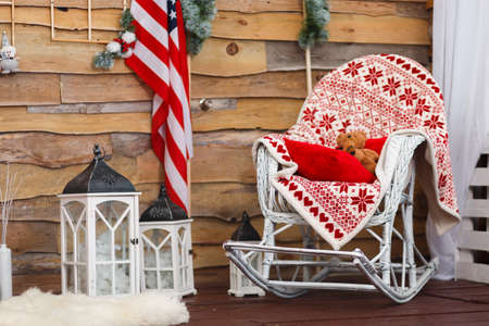Decorated house in Christmas style. The rocking chair is covered with a red rug in which there is a teddy bear, decorated with fir tree branches and an American flag on wooden walls. Indoors.の写真素材
