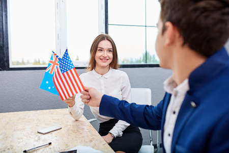 Office workers, a guy and a girl, are holding flags of america and australia. Inside the office.