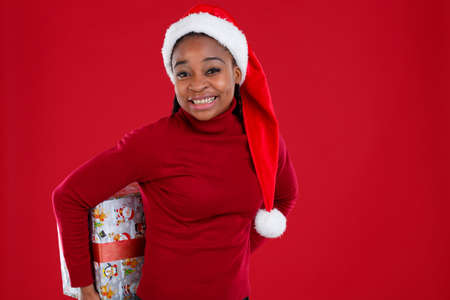 Smiling African-American girl in Santas hat, holding behind the back a gift box and smiling broadly.の写真素材