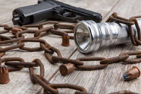 A rusty chain, bullets, a gun and a silver flashlight lie on a wooden background