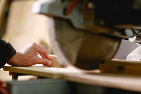 Hands of the employee in focus hold the board on a circular saw for cutting on a blurred background....