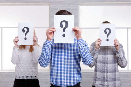 Two guys and a girl stand side by side close their faces with sheets with question marks. The guy in...