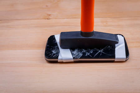 A broken phone with an insulating tape from above and below is a hammer on a wooden floor. Top view of a closeupの写真素材