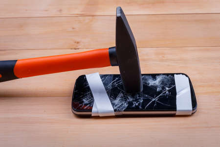 The broken phone is rewound with a white tape at the bottom and on top of which stands a hammer on a wooden floor. Top view of a closeupの写真素材