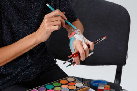 A close-up smeared hand make-up artist in paint, holds a brush, and a color palette of paints, selects the color of the paint by doing strokes on the hand.の写真素材