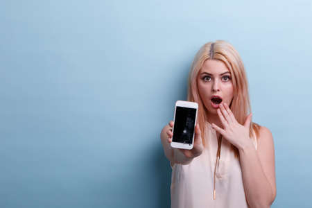 A girl very surprised shows a broken phone in an outstretched hand on a blue backgroundの写真素材