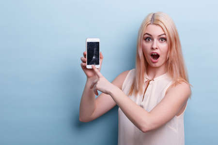 A girl in shock shows at a broken phone and looks directly at a blue backgroundの写真素材