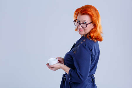 A woman stands sideways and holds a cup in her hands on a gray background