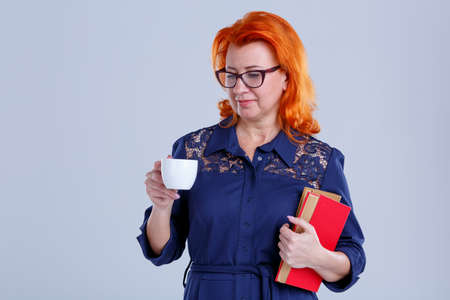 A woman stands with books and looks at the cup in her hand on a gray background