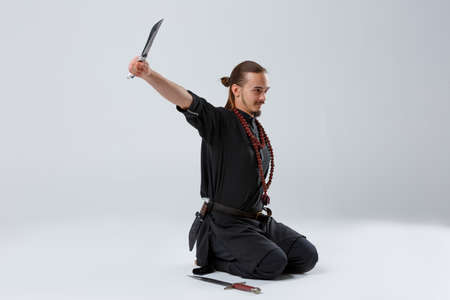 A ninja man sits kneeling with a knife in his outstretched arm on a gray background
