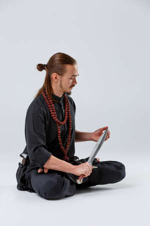A ninja man sits in a lotus pose with a sword in his hand on a gray backgroundの写真素材
