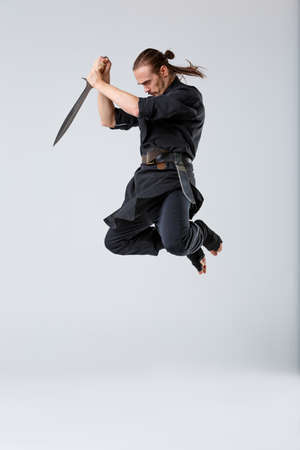 A ninja man in a jump holds the sword with both hands pointing down on a gray backgroundの写真素材