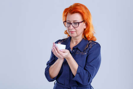 A woman with glasses holds a mug in her hands and looks at it against a gray backgroundの写真素材