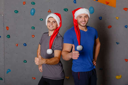 Two sports guy in santa hats smile and show thumb up. Against the wall for rock climbing.の写真素材