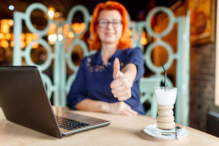 Woman not in focus shows thumb up at table in cafe with laptop