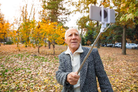 An elderly man in an autumn park and holds a monopod with a mobile phone. Outdoors on the street.の写真素材