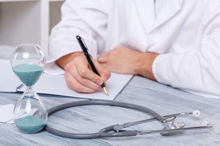 Doctor's hands make notes in a notebook sitting on a table on which an hourglass and a stethoscope close-upの写真素材