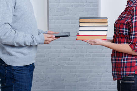 Two young people stand opposite each other and holds the pile of books, and modern tablet.