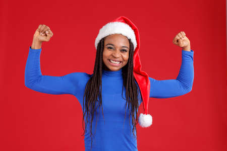 A happy african american girl in blue golf and a Christmas hat shows the biceps of her hands and is smiling.の写真素材
