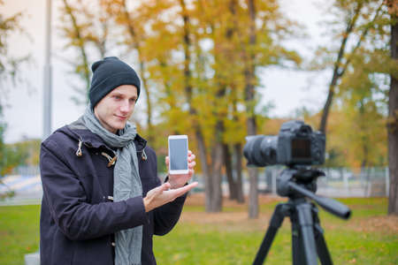 A guy takes pictures of himself on a camera on a tripod showing off a mobile phone. Outdoors.の写真素材