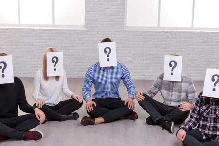 A group of five people sit on the floor, on their faces are sheets with a question mark.の写真素材