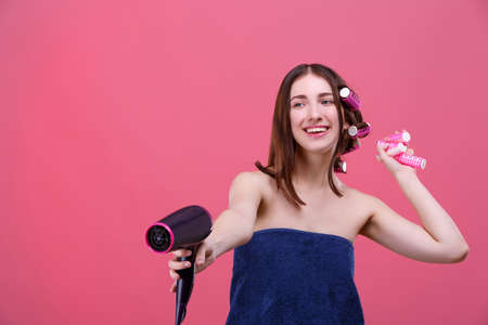 A happy girl, in a blue bath towel, with curlers, holds a hair dryer in front of her and laughs.